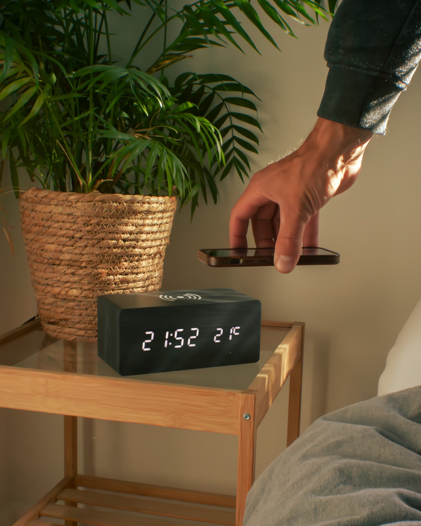 Person placing a smartphone on a digital clock with plant and wooden nightstand in the background